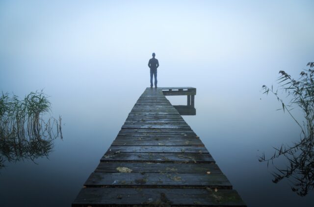 Man standing on a jetty at a lake during a foggy, gray morning.
