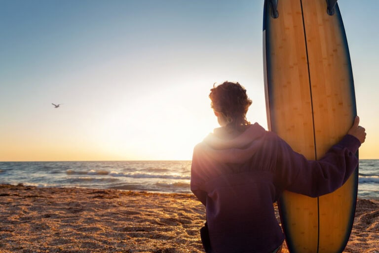 Surfer with surfboard sitting on the beach at sunset